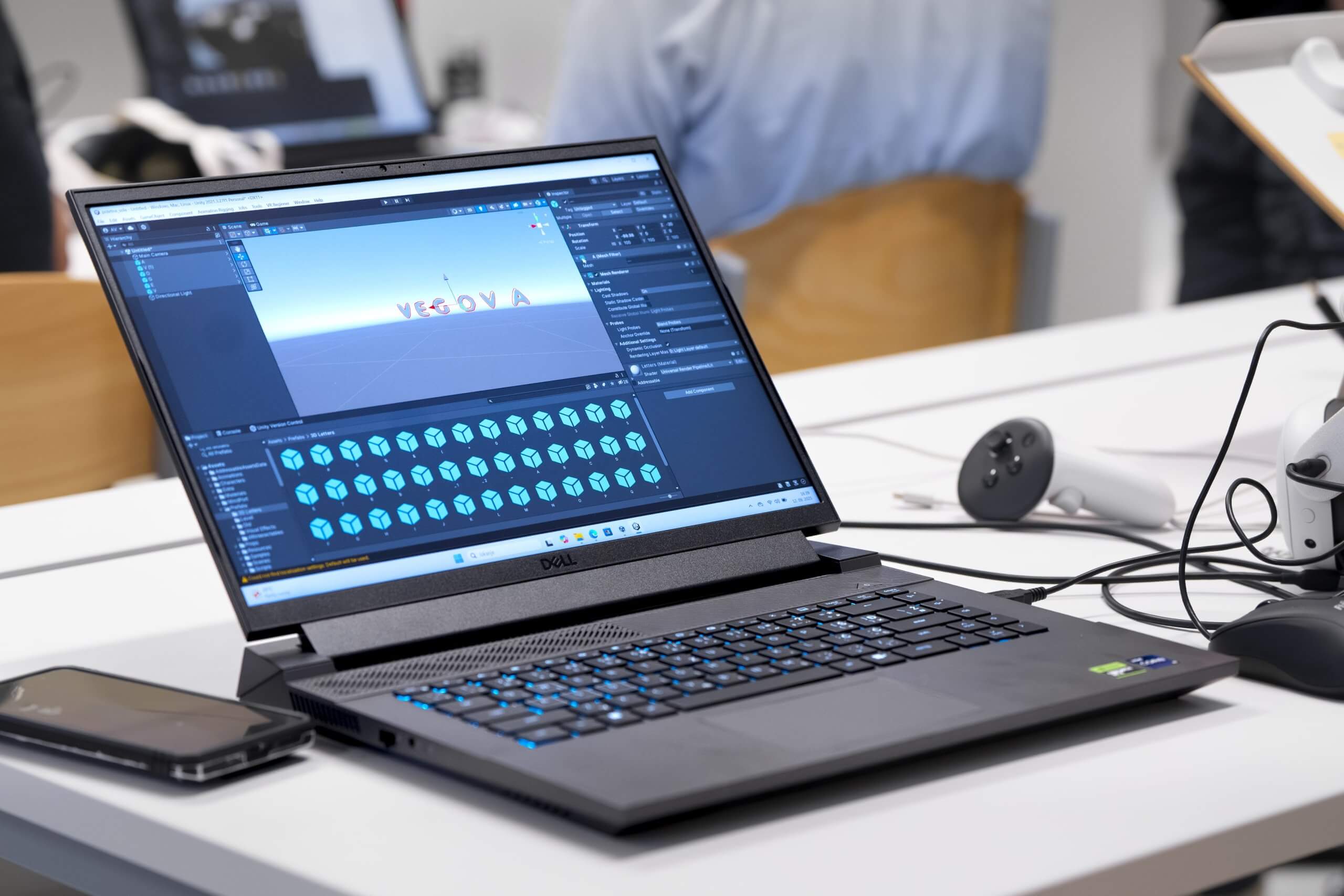 Unity na Vegovi Na prenosnem računalniku je odprt program Unity, kjer je viden napis VEGOVA. Na mizi je tudi mobilni telefon, krmilnik in očala za navidezno resničnost.