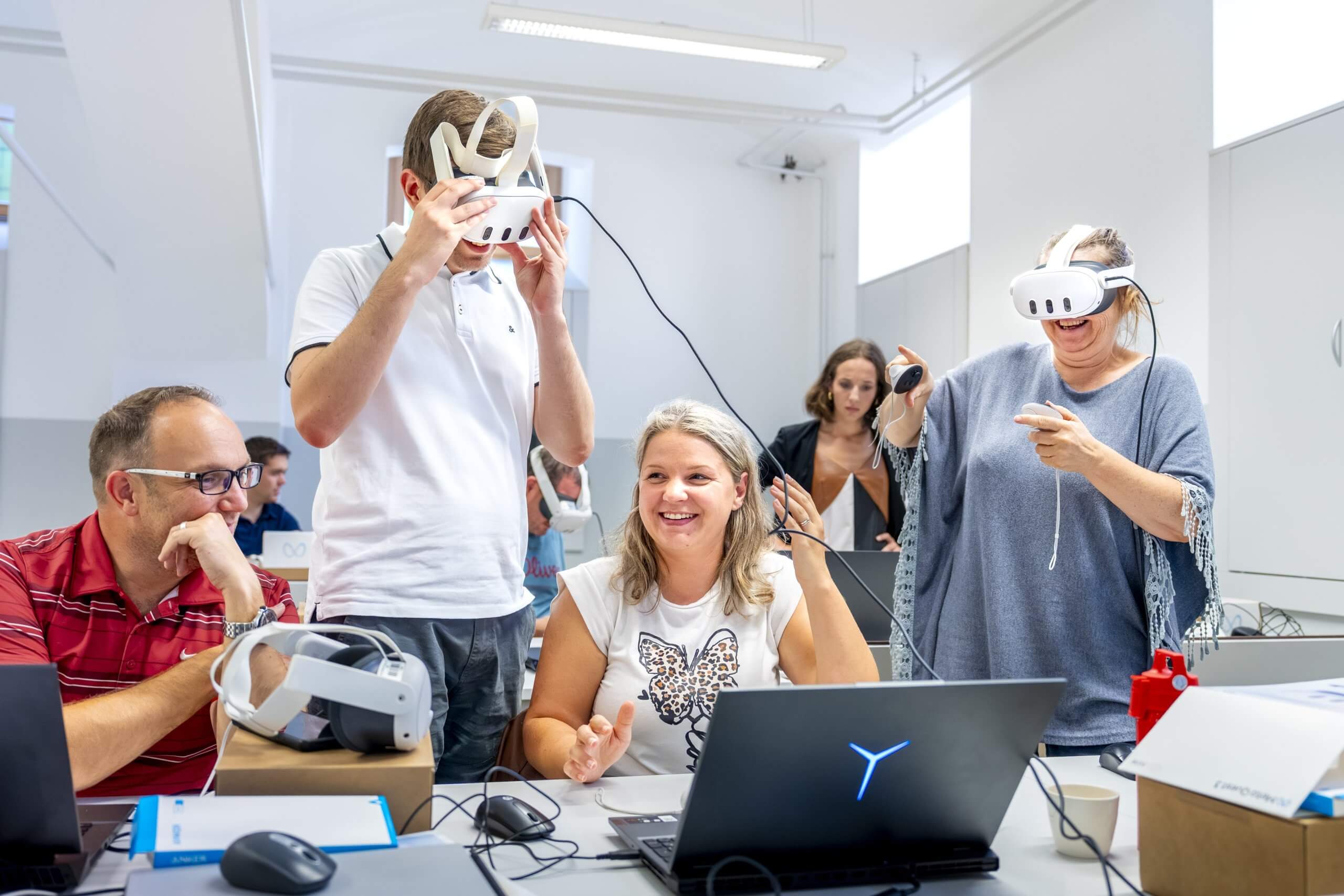 Testiranje izdelkov na očalih Udeleženci delavnice na očalih za navidezno resničnost testirajo izdelke, ki so jih pripravili v programu Unity. Moški in ženska sedita ob mizi z računalnikom, za njima stojita drug moški in ženska, ki imata na glavah očala in v rokah krmilnike.