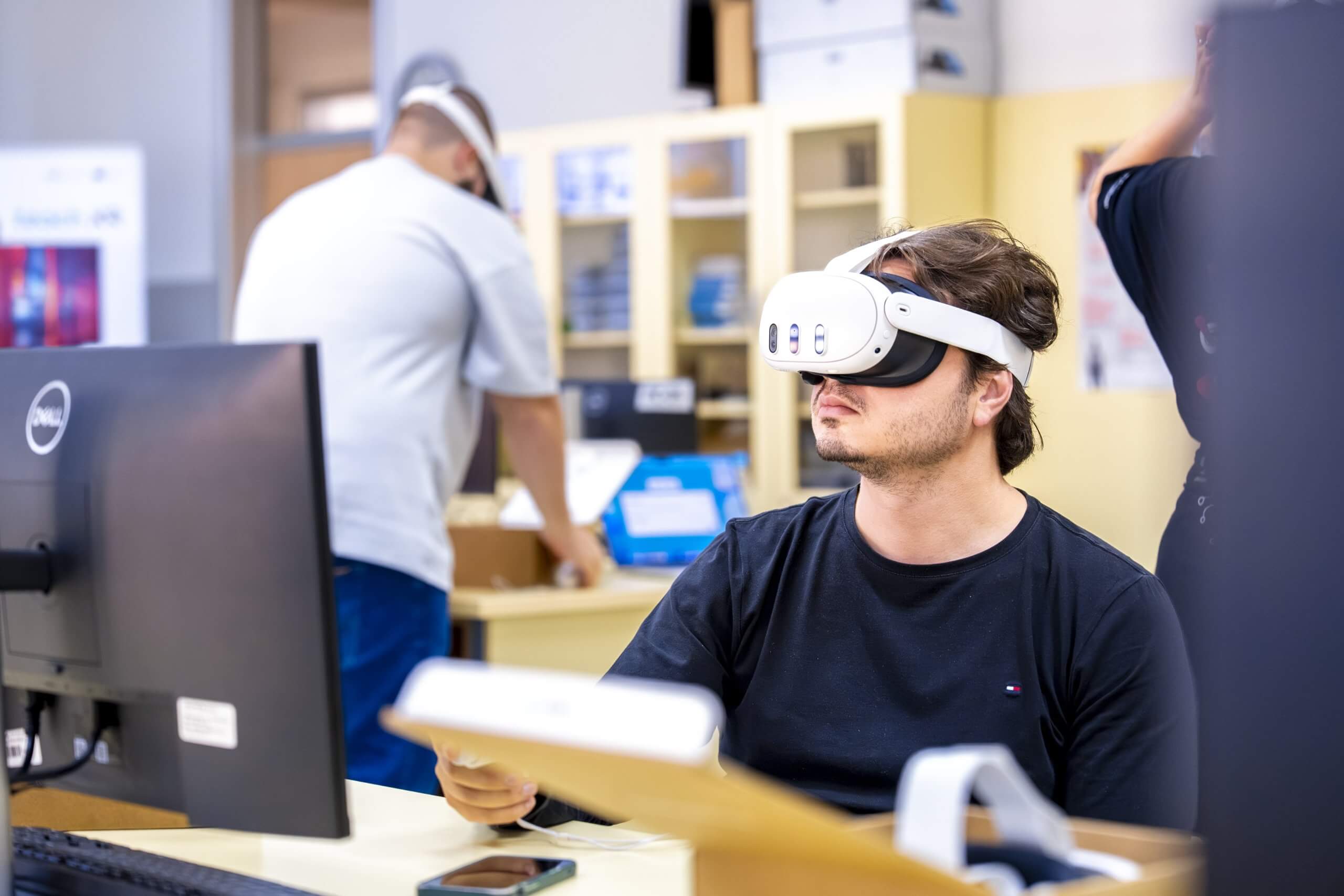 Moški z VR očali na glavi in krmilnikom v roki sedi na stolu in gleda vsebine navidezne resničnosti, v ozadju poteka predavanje.