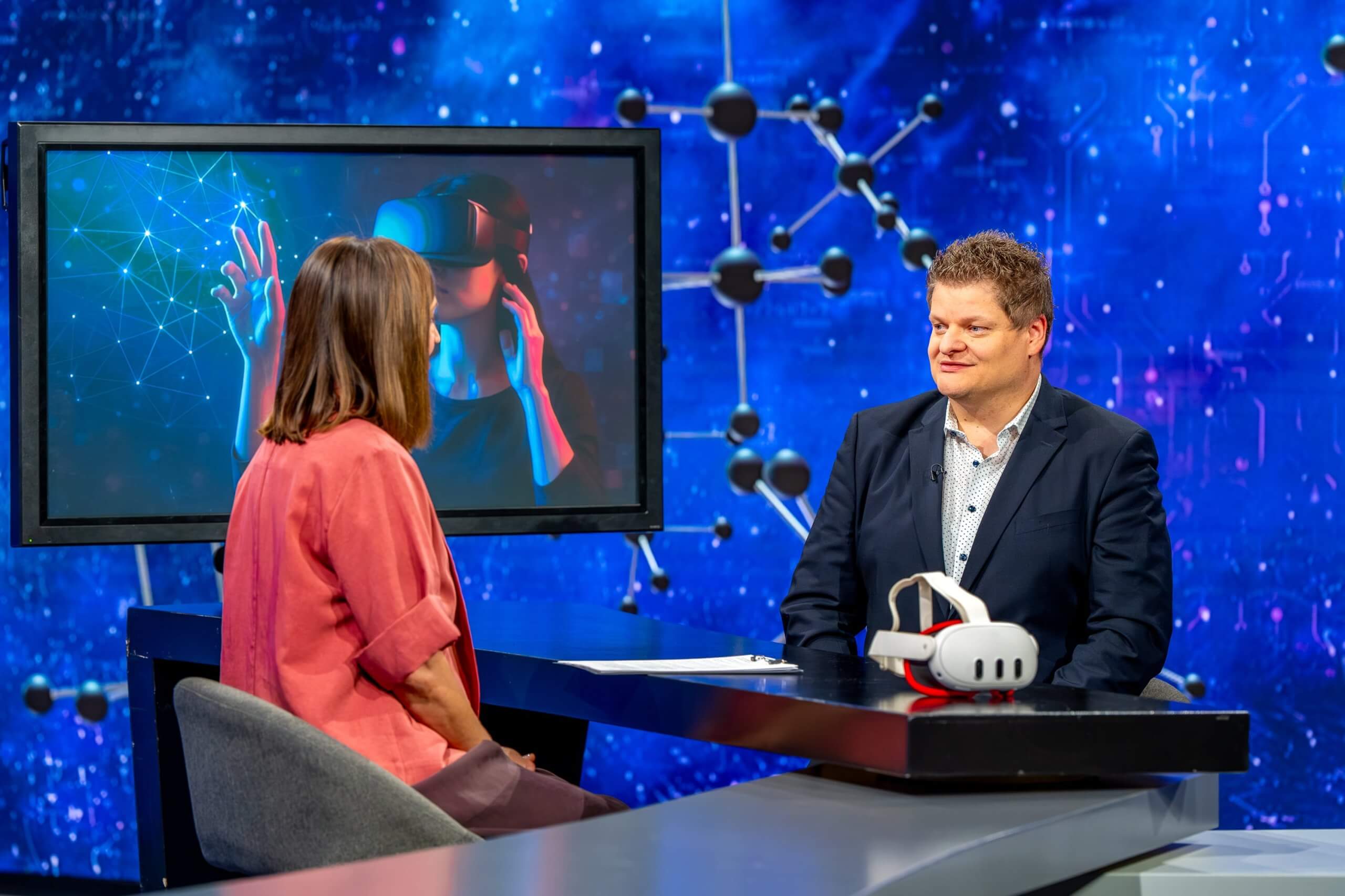 Ugriznimo Znanost Za televizijskim pultom na levi sedi voditeljica, ki se pogovarja z gostom (Klemnom Pečnikom) na desni. Na pultu so očala za navidezno resničnost v ozadju je televizija in strukture kemijskih formul.