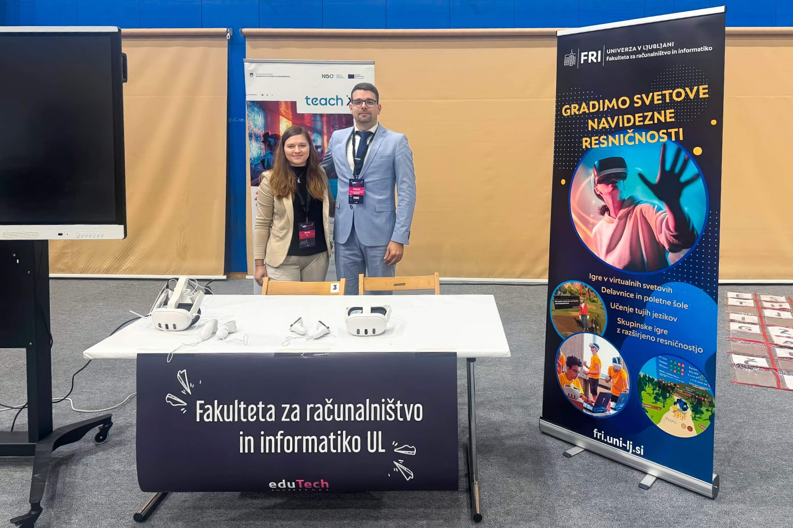 edutech konferenca Za mizo, na kateri so postavljena VR očala, stojita moški in ženska. Za njima je postavljen plakat za TeachXR, na desni pa plakat za izzobraževanje LingoVerse.