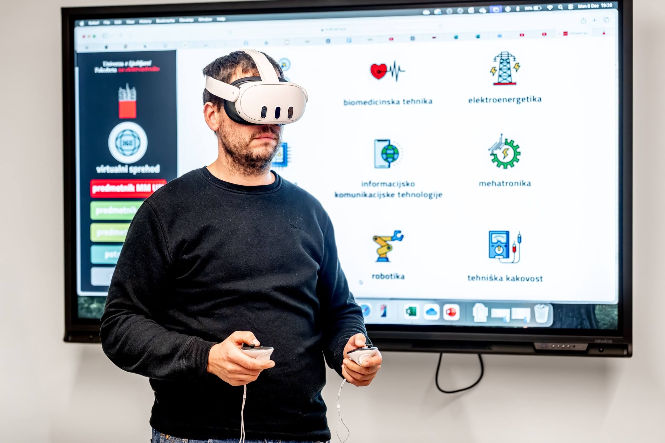 Udeleženec izobraževanja z očai za navidezno resničnost na glavi in krmilniki v rokah, stoji pred interaktivno tablo, z vidno vsebino.